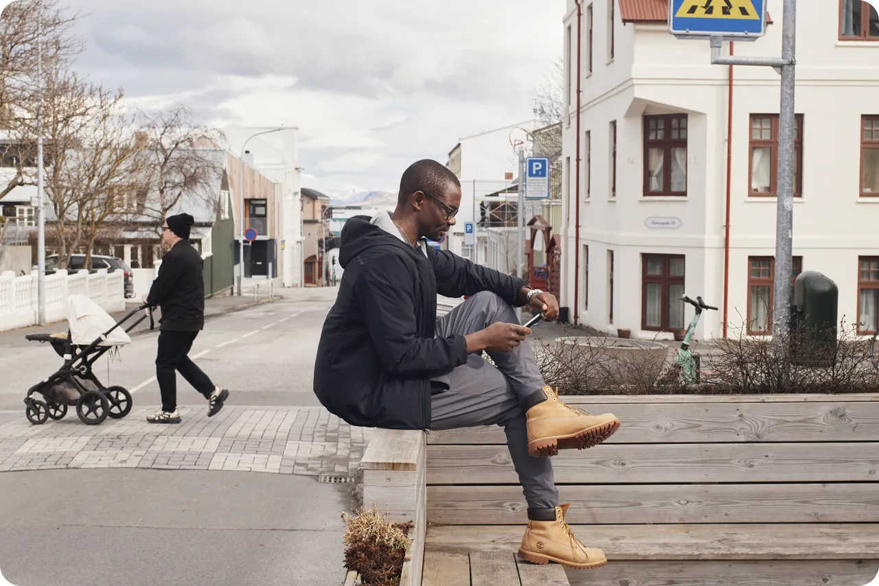 Someone sits on a wooden bench by the street, looking at a phone